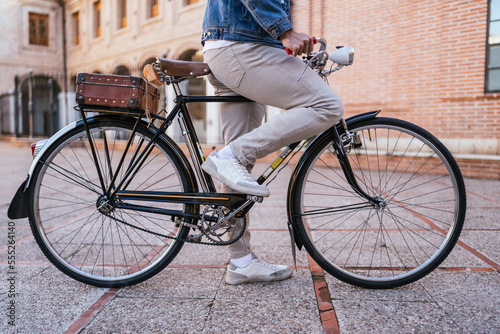 Cropped profile image of a young man in a denim jacket sitting atop his vintage classic bicycle with his foot on the pedal in an attempt to start the ride.