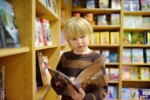A preteen boy leafing through a book while standing at a bookshelf in a school library or bookstore.