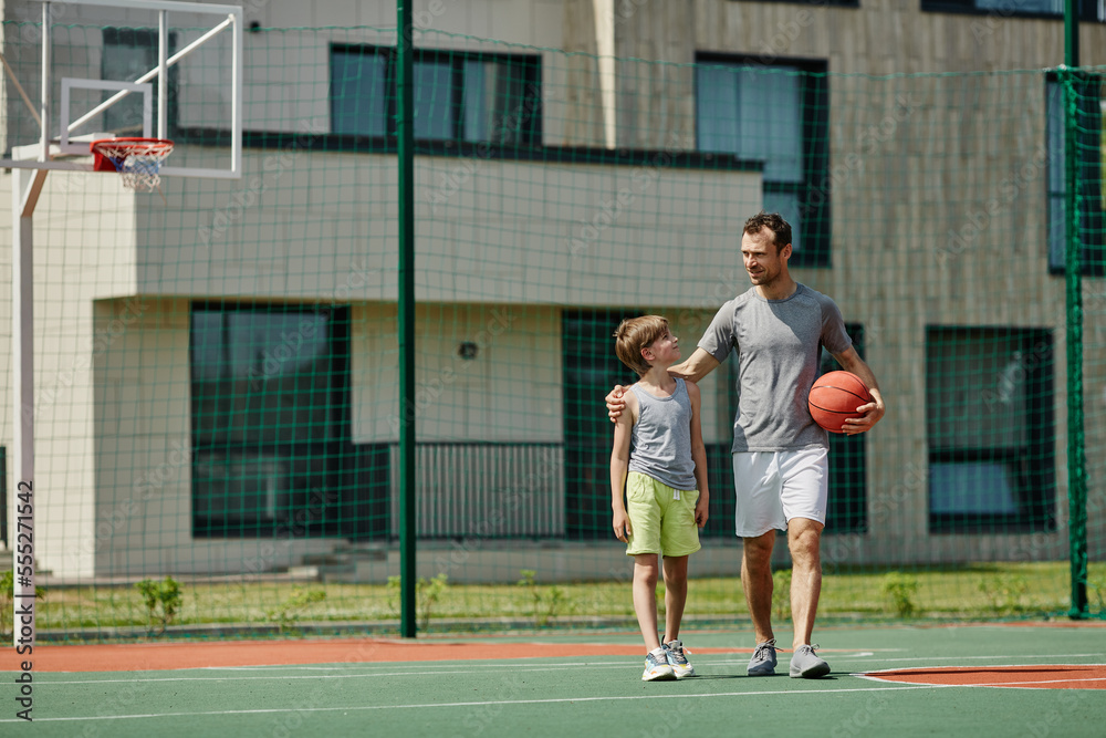 Full length portrait of happy father and son playing basketball together and standing on court in sunlight, copy space