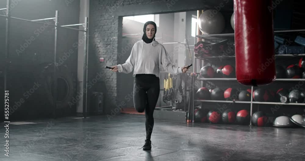 Attractive active sporty Arab woman in hijab training in gym jumping with skipping rope. Beautiful fit muslim female in leggings and hoodie working out doing sports keeping healthy lifestyle.