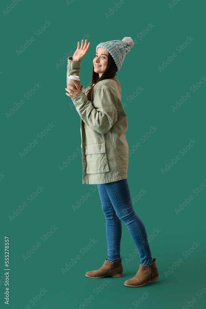 Young woman in warm hat with takeaway cup of coffee waving hand on green background Young woman in warm hat with takeaway cup of coffee waving hand on green background