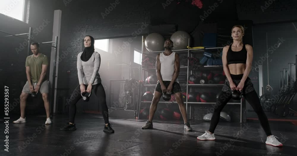Group training session of 4 sporty people. Attractive fit skinny powerful caucasian, African and Arab muslim men and women training legs doing plie squats with bells weights in crossfit gym.
