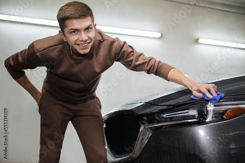 Car wash and cleaning at professional auto service station. Caucasian young male worker cleaning car headlights with blue microfiber cloth.