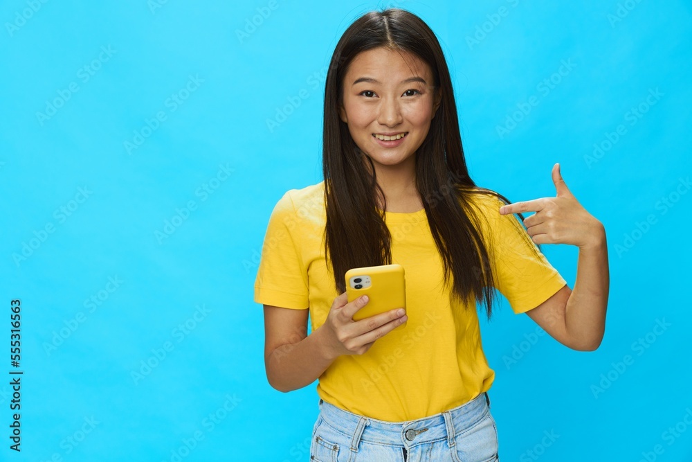 Happy Asian woman holding a phone with a yellow case on a blue background in a yellow T-shirt with a smile and teeth pointing her finger at Happy Asian woman holding a phone with a yellow case on a blue background in a yellow T-shirt with a smile and teeth pointing her finger at