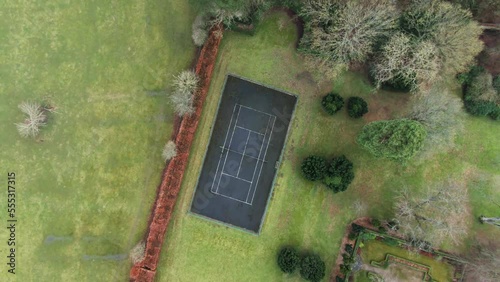 Top down drone view of private asphalt tennis court in garden surrounded by trees, grass and fields