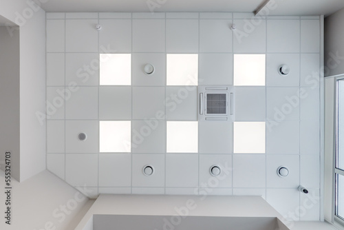cassette stretched or suspended ceiling with square halogen spots lamps and drywall construction with fire alarm and ventilation in empty room in house or office. Looking up view