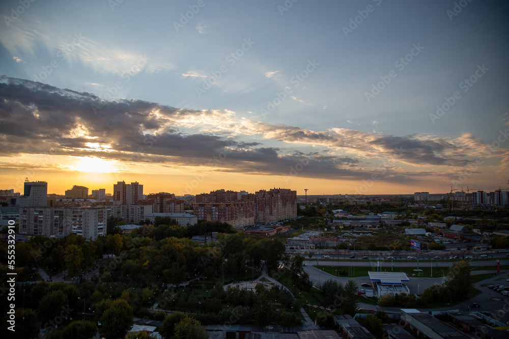 Sunset breaks through big clouds in sky and illuminates busy city. Multi-storey buildings stand surrounded by green trees in evening