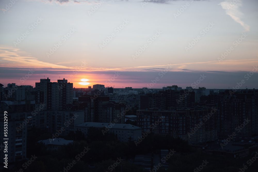 Sunset signals about upcoming night and illuminates high-rise city buildings. Streets become darker and trees silhouettes surround residential area