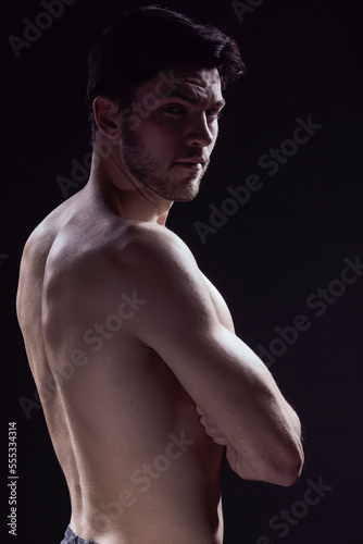 Natural Portrait of Confident Thinking Caucasian Bodybuilder Athlete Man Posing With Naked Torso Looking Backwards Against Dark