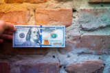 A hand holds a hundred dollar bill against the background of an old brick wall close-up. Old crumbling red brick wall and cash, us dollar