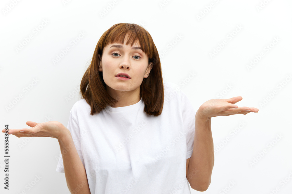 a pleasant, beautiful, attractive woman stands on a white background in a white T-shirt and looking at the camera, she spreads her arms in surprise to the sides a pleasant, beautiful, attractive woman stands on a white background in a white T-shirt and looking at the camera, she spreads her arms in surprise to the sides