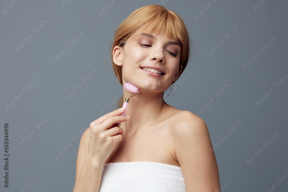 a happy woman with perfect skin stands on a gray background and, covering her eyes with pleasure, does a facial massage with a pink roller, smiling pleasantly. Close horizontal photo a happy woman with perfect skin stands on a gray background and, covering her eyes with pleasure, does a facial massage with a pink roller, smiling pleasantly. Close horizontal photo