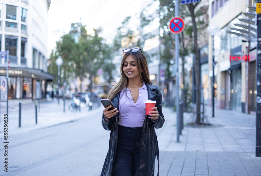 Woman reads a message on the smartphone. In the left hand she holds a coffee mug. Modern, confident, outdoors, street, urban place, center of the city and summer.