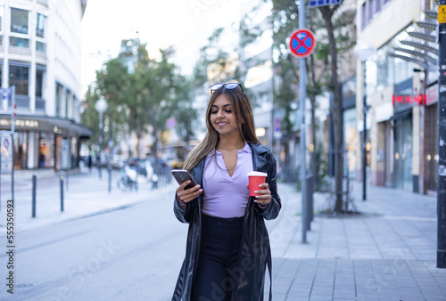Woman reads a message on the smartphone. In the left hand she holds a coffee mug. Modern, confident, outdoors, street, urban place, center of the city and summer. © Westlight