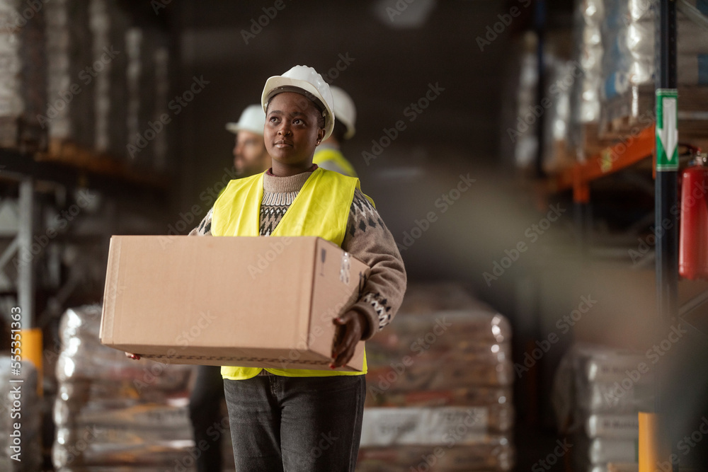 A female warehouse worker wearing protective clothing is carrying a box in a large warehouse.