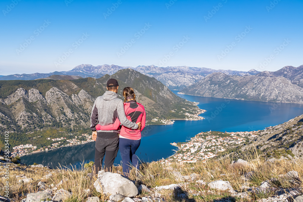 Loving couple hugging with panoramic view of Kotor bay in sunny summer, Adriatic Mediterranean Sea, Montenegro, Balkan, Europe. Fjord winding along coastal towns. Lovcen and Orjen mountain range