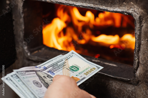 American money in hand of a person near opened door of solid fuel boiler, close up. Expensive heating of the house, warmth and comfort at home due to large expenses photo