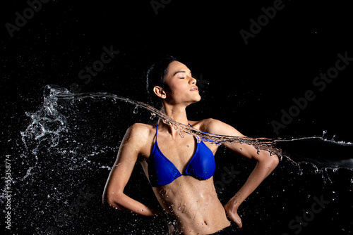 Tanned skin Asian woman in bikini poses in aqua studio. Splash Drops of water spread to body. Fun emotion female girl on water attack fluttering and stop motion freeze shot, black background isolated