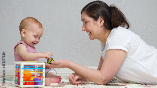 Mother plays with baby daughter against white wall. Child with excited expression enjoys playing with toys and smiles demonstrating love to mother