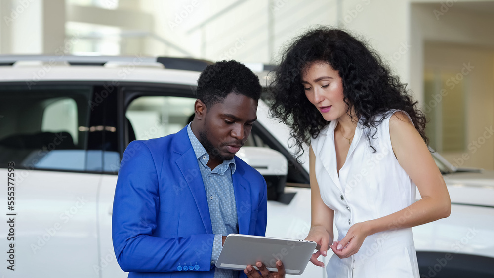 African American male car dealer presents car to female customer in automobile salon. Black salesman shows characteristics on tablet and describes details African American male car dealer presents car to female customer in automobile salon. Black salesman shows characteristics on tablet and describes details