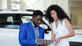 African American male car dealer presents car to female customer in automobile salon. Black salesman shows characteristics on tablet and describes details