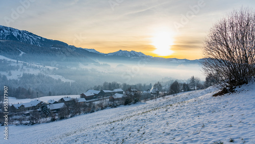 Panorama mit Waldrand, Dorf und Schnee auf Wiese und im Hintergrund die Berge des Bregenzerwaldes. verschneite Landschaft, Nachmittagssonne vor Sonnenuntergang, beleuchtete Landschaft im Winter, 