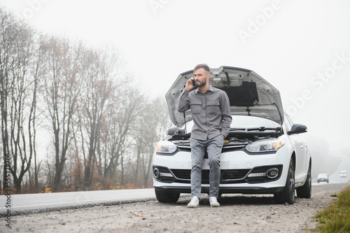 Sad driver calling car service, opening hood, having engine problem standing near broken car on the road. Car breakdown concept