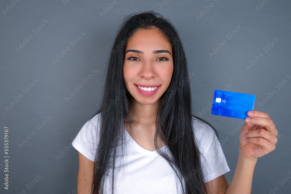 Young beautiful hispanic woman wearing white t-shirt over gray studio background, smiling and presenting a blue credit card. Online shopping and finances concept. Young beautiful hispanic woman wearing white t-shirt over gray studio background, smiling and presenting a blue credit card. Online shopping and finances concept.