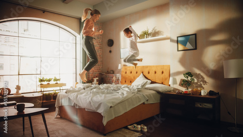 Mother and Small Daughter Having Fun at Home. Cheerfully Playing, Jumping on a Bed in Stylish Loft Apartment. Happy Childhood, Parenthood and Motherhood Memories. Wide Angle Footage.