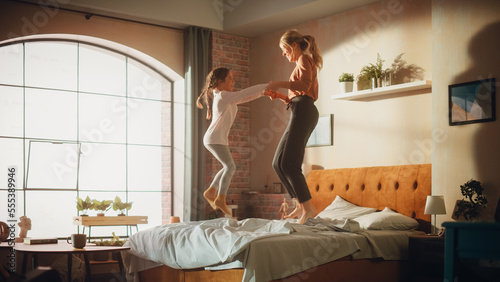 Adorable Little Girl Having Fun at Home with Young Beautiful Mother. Family Playing at Home, Cheerfully Jumping and Falling onto a Bed in Stylish Loft Apartment with Big Window. Happy Childhood. photo