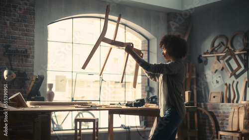 Portrait of a Young Beautiful Carpenter Checking a Blueprint and Starting to Assemble a Wooden Chair. Professional Furniture Designer Working in a Studio in Loft Space with Tools on the Walls. © Gorodenkoff