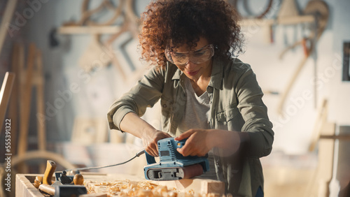 Fotografia Multiethnic Woman Carpenter Wearing Protective Safety Glasses and Using Electric Belt Sander to Grind a Wood Block