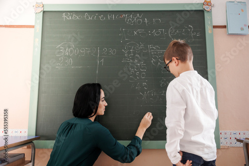 Maestra scrive con il gesso alla lavagna delle formule matematiche mentre lo studente guarda attento photo