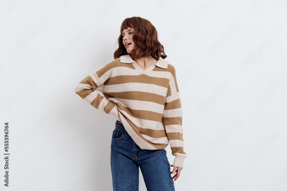 horizontal photo on a light background of a cute, pleasant woman in a stylish striped sweater correcting it on one side on the belt while standing in blue jeans horizontal photo on a light background of a cute, pleasant woman in a stylish striped sweater correcting it on one side on the belt while standing in blue jeans