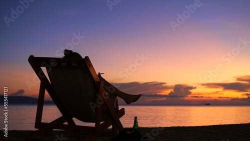 On a sunset beach, a girl sits in an armchair smoking a cigarette and drinking beer while enjoying her vacation. Holidays at sea in the tropics. The concept of tourism in Asia and the Caribbean.
