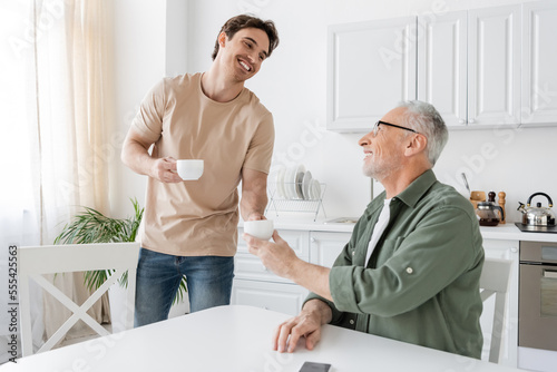 young and happy guy giving coffee cup to smiling grey haired father sitting in kitchen