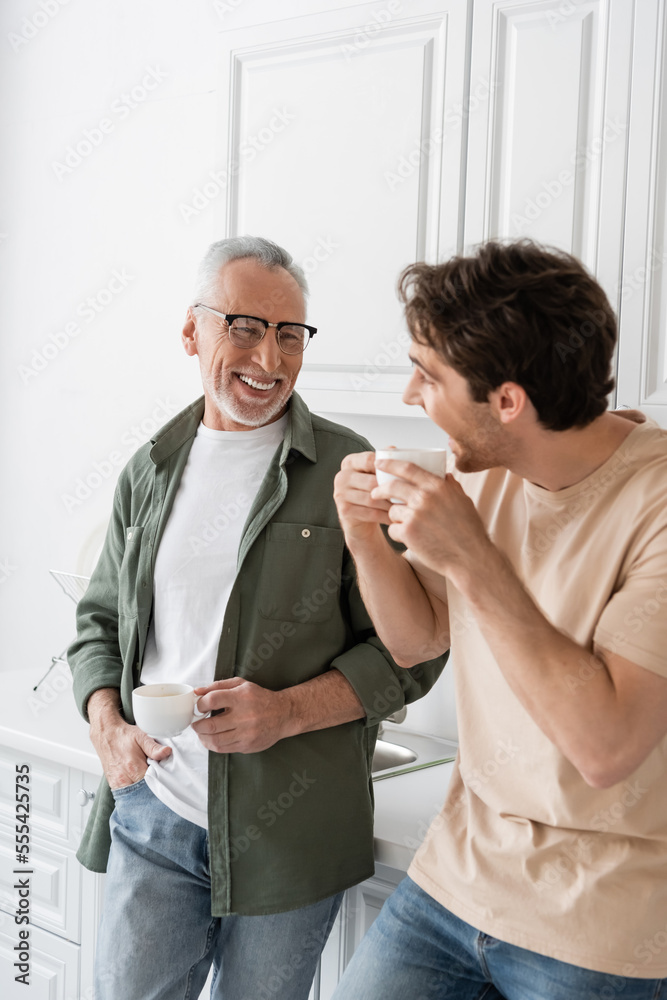 Fototapeta premium young man holding coffee cup and looking at cheerful dad during conversation in kitchen