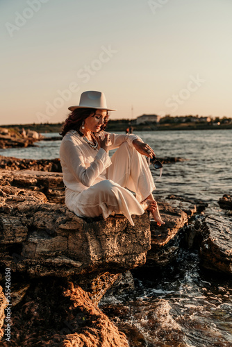 A woman in a white pantsuit and hat is standing on the beach enjoying the sea. Happy summer holidays