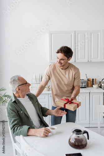 smiling man presenting fathers day gift to pleased dad sitting in kitchen with coffee cup