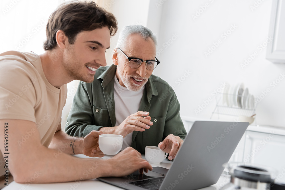 cheerful mature man pointing at laptop near young son and coffee cups in kitchen cheerful mature man pointing at laptop near young son and coffee cups in kitchen