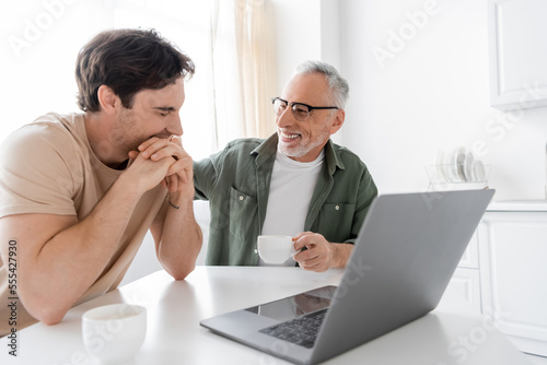 cheerful grey haired man with coffee cup looking at young son smiling with closed eyes near laptop
