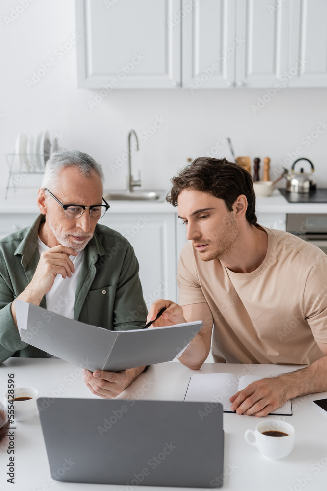 young man pointing with hand near thoughtful dad holding folder with documents while sitting at laptop young man pointing with hand near thoughtful dad holding folder with documents while sitting at laptop