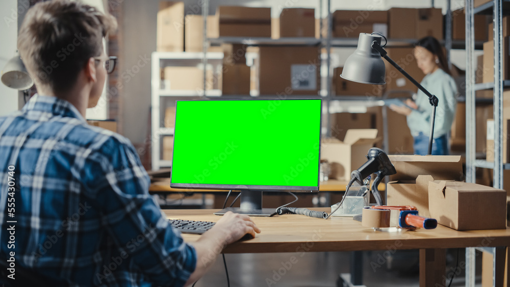 Young Man Using Desktop Computer with a Green Screen Chromakey Mock Up Display. Distribution Manager and Employee Working in Distribution Warehouse with Cardboard Boxes. Over the Shoulder Footage. Young Man Using Desktop Computer with a Green Screen Chromakey Mock Up Display. Distribution Manager and Employee Working in Distribution Warehouse with Cardboard Boxes. Over the Shoulder Footage.