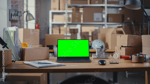 Desktop Computer Monitor Standing on a Table with a Green Screen Chromakey Mock Up Display. Small Business Warehouse in the Background. Desk with Cardboard Boxes