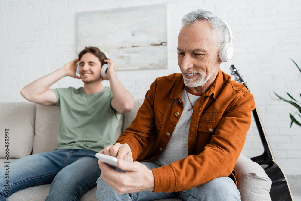 mature and happy man in wireless headphones using smartphone near son listening music on blurred background