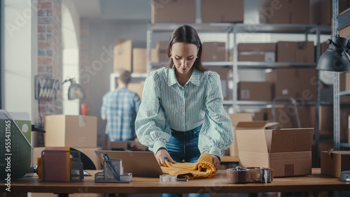Small Business Owner of an Online Store Works on Laptop Computer while Leaning On Her Desk in Warehouse. Female Employee Packing a Stylish Yellow Jumper in the Room with Shelves Full of Parcels. © Gorodenkoff