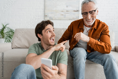 excited man pointing at cheerful mature dad while holding blurred mobile phone