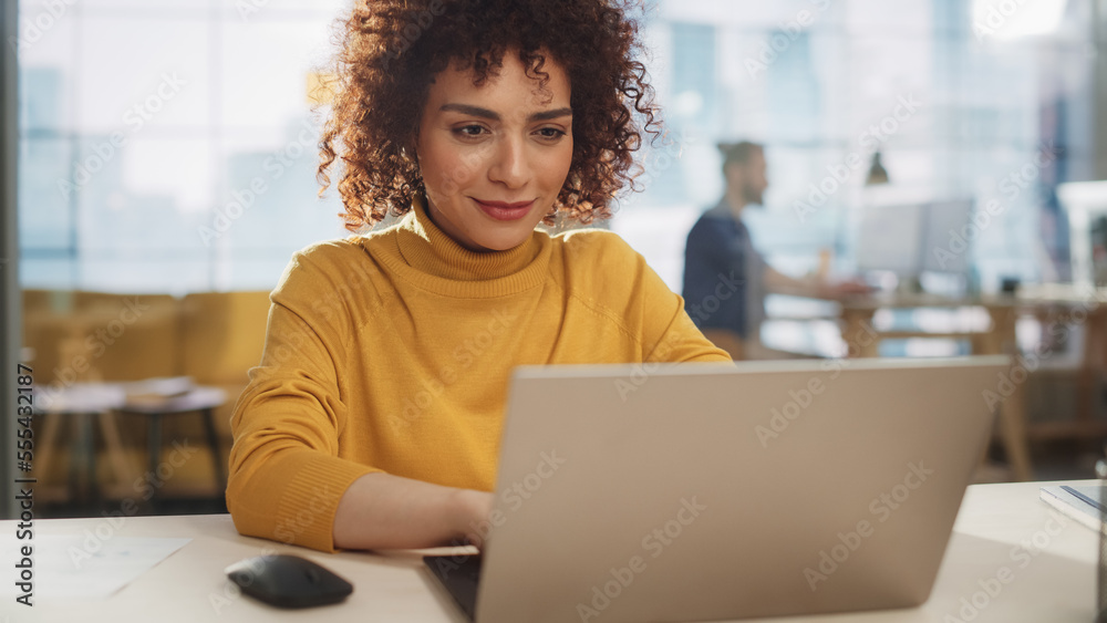 Close Up Portrait of a Happy Middle Eastern Female Sitting at a Desk in Creative Agency. Young Stylish Female with Curly Hair Using Laptop Computer in Marketing Agency. Close Up Portrait of a Happy Middle Eastern Female Sitting at a Desk in Creative Agency. Young Stylish Female with Curly Hair Using Laptop Computer in Marketing Agency.