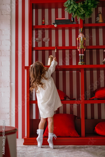 Cute little curly-haired girl in dress on tiptoe reaches for xmas bell on shelf photo