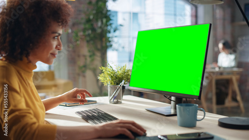 Portrait of an Excited Multiethnic Specialist Working in Creative Agency. Stylish Expressive Female in Yellow Jumper Using Desktop Computer with Green Screen Mock Up Display.